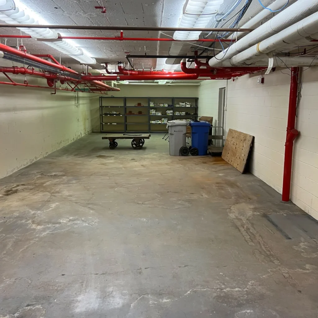 Large emptied basement space with red and white pipes overhead and a clean floor