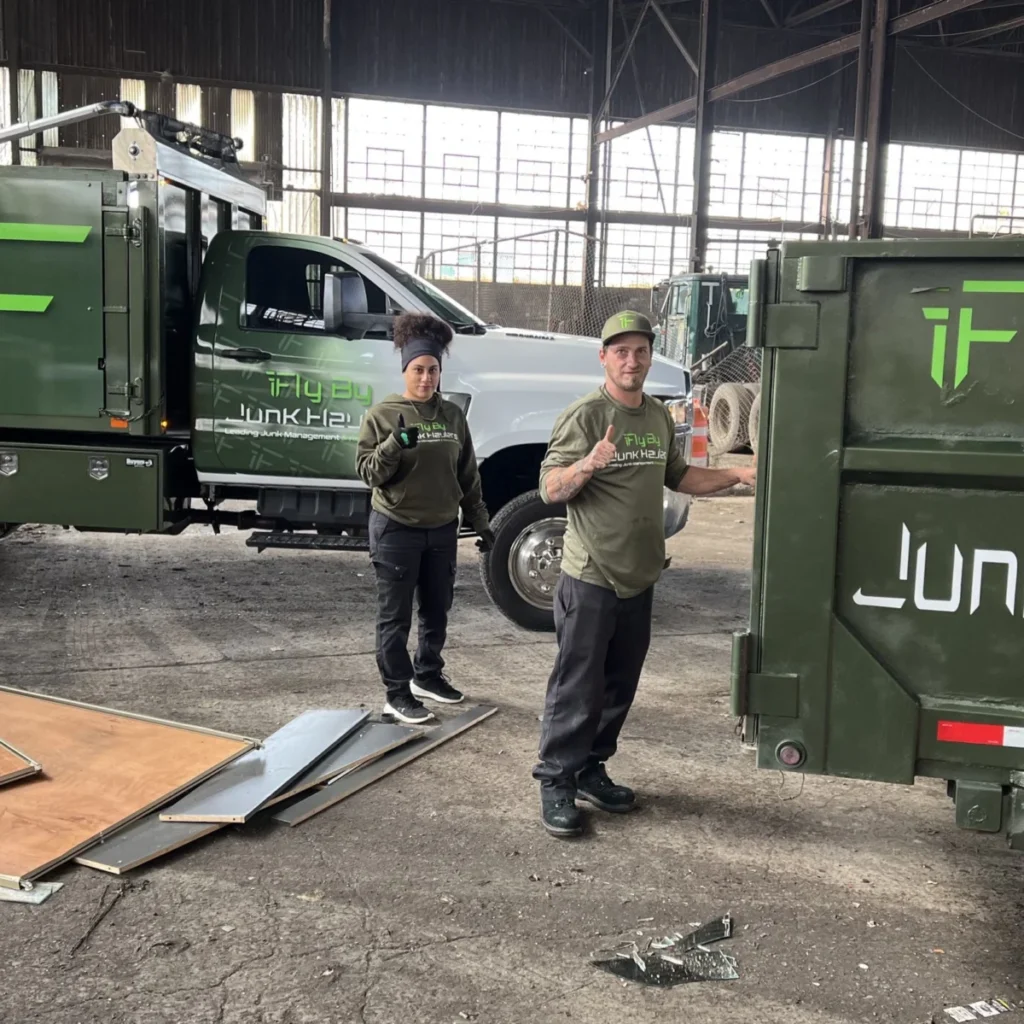 Two Fly By Junk Haulers crew members beside a hauling truck and a green dump trailer inside a large commercial building