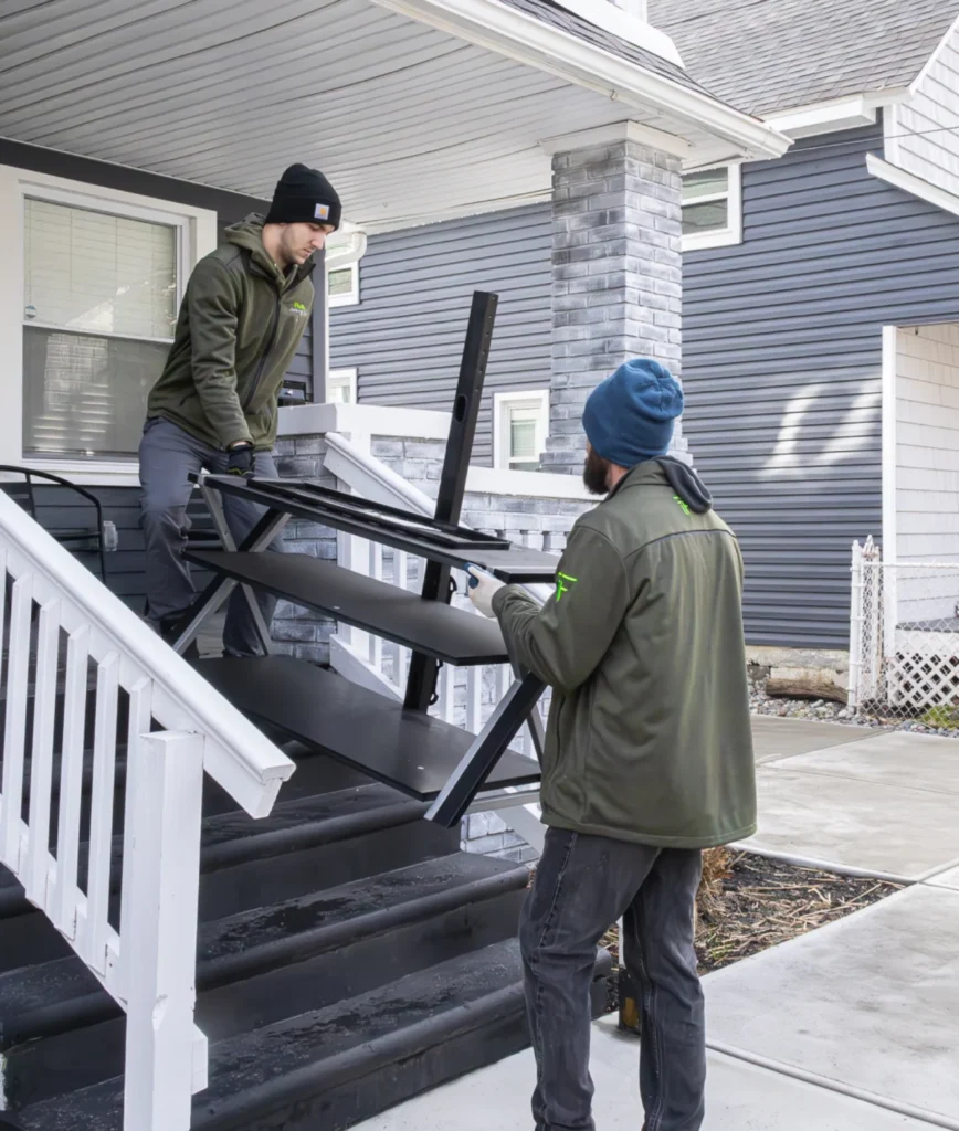 Two Fly By Junk Haulers crew members carrying a metal shelf down porch steps