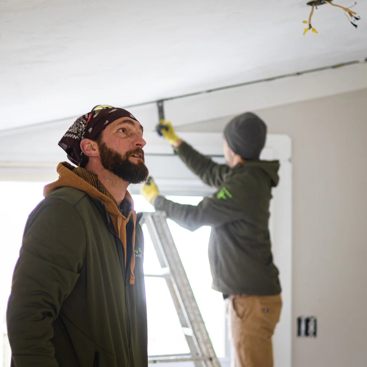 Two Fly By Junk Haulers crew members working on a ceiling in a bright empty room, one on a ladder