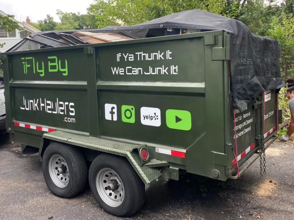 Green Fly By Junk Haulers dump trailer with a tarp over the load, parked in a driveway