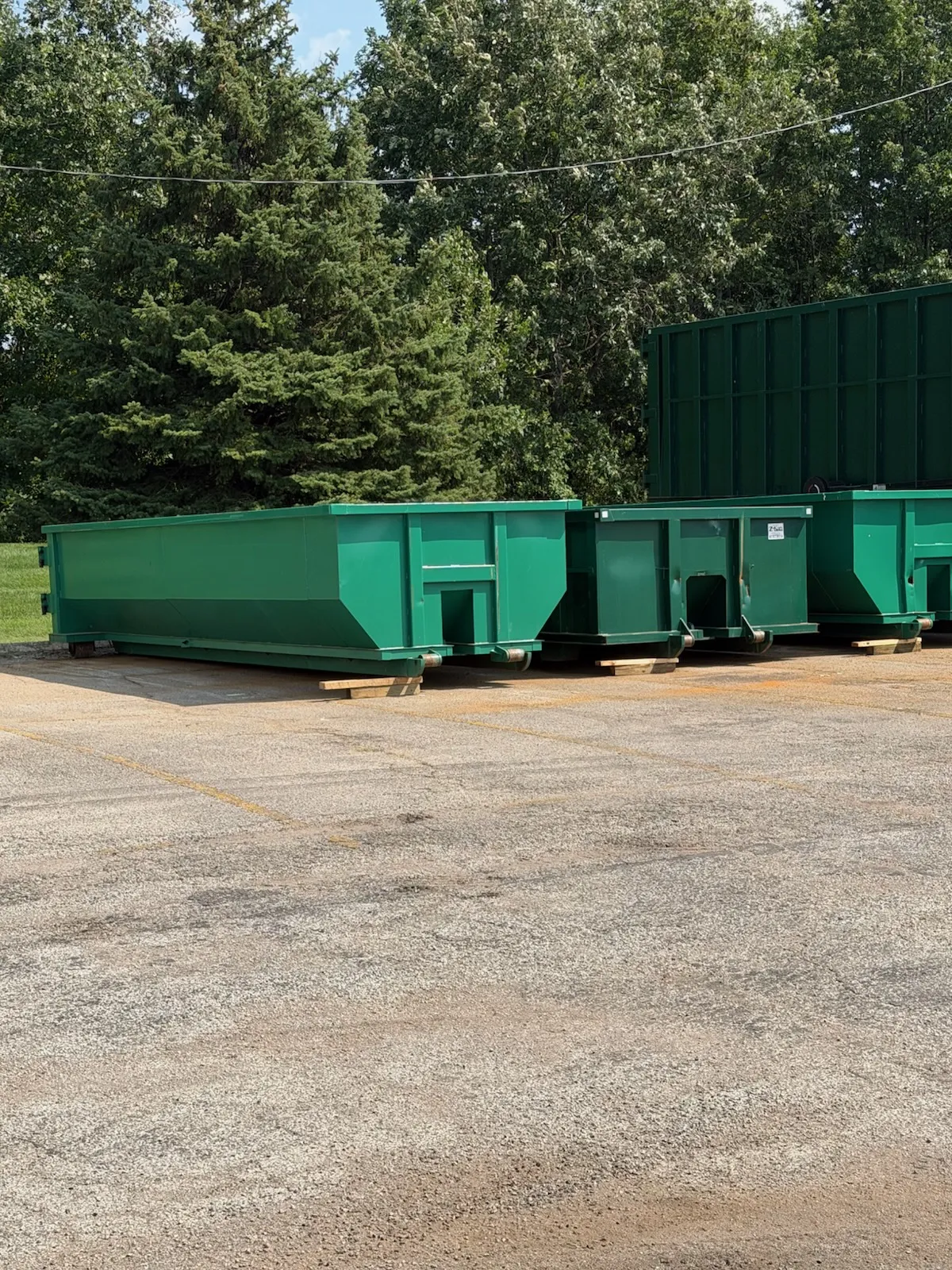 Green roll-off dumpsters parked on a gravel lot with trees behind
