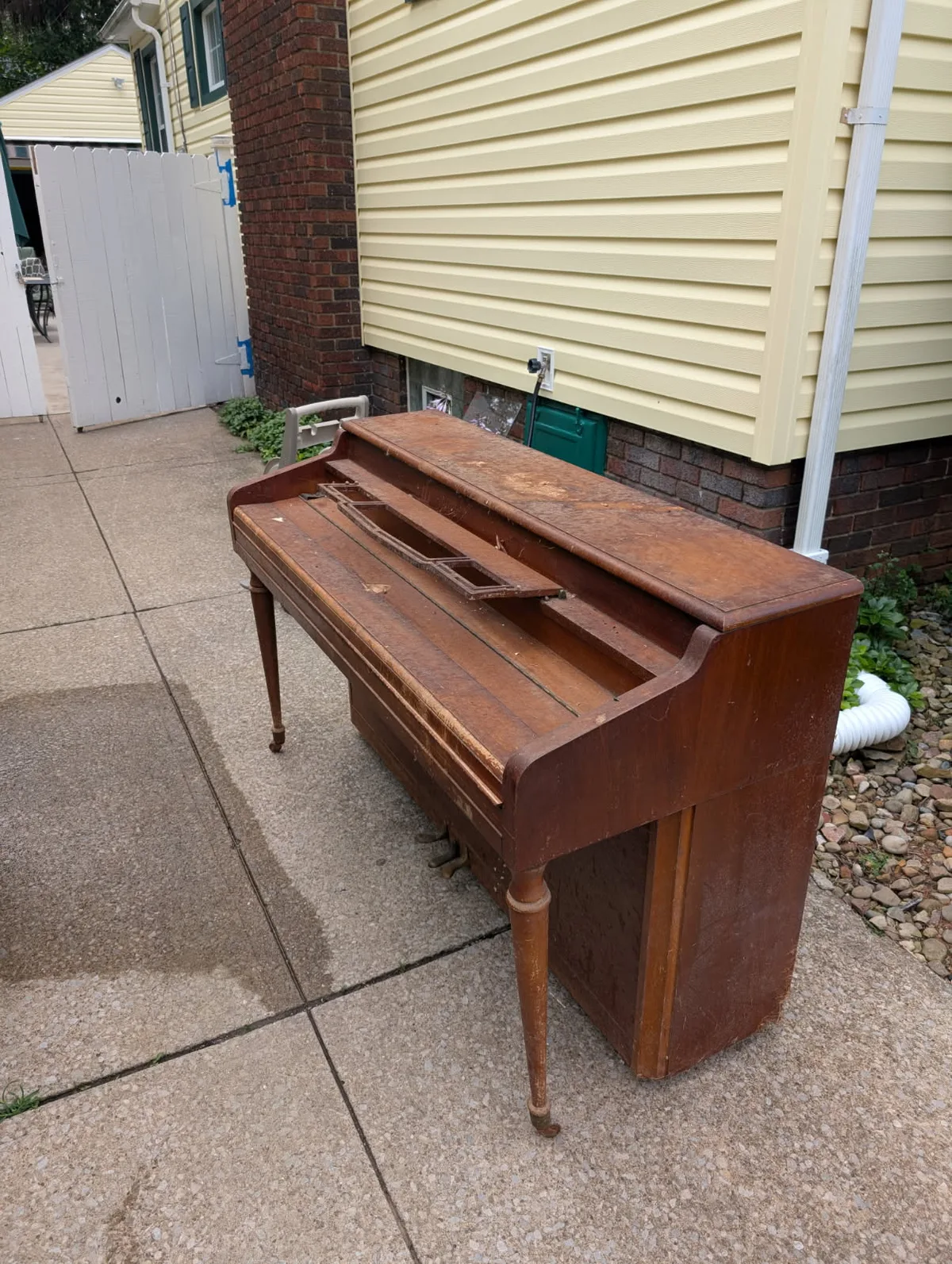 Old spinet piano sitting on a driveway beside a house