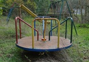 Playground in overgrown garden of an abandoned house. Rusty merry-go-round swings and slide.